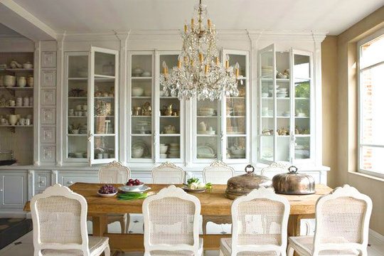 Dining room in Belgium with white drawers and cabinets with glass fronts, cane back chairs, a chandelier, farmhouse style wood table 