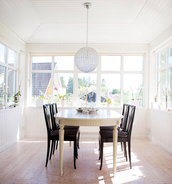 Bright white sunroom with an oval Gustavian white table, dark wood dining chairs, and a crystal ball chandelier