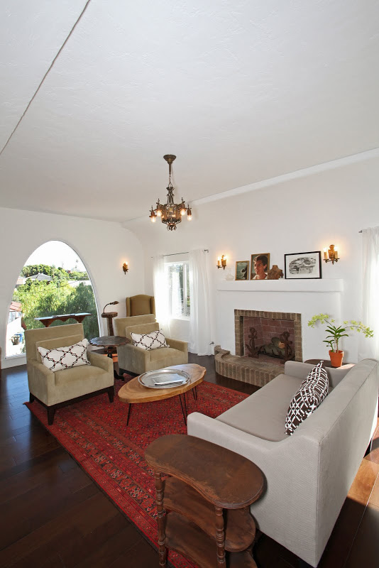 Alternative view of the living room with arched window, club chairs and a grey sofa with cococozy pillows and throws, dark wood floors, a red Moroccan rug, a brick fireplace and a chandelier