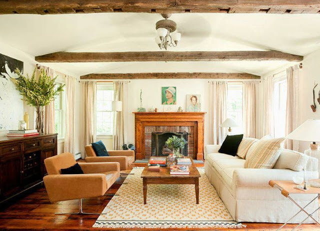 Living room in a farmhouse with exposed beam ceiling, a fireplace with a wood mantel, wood floor, a long white sofa, a reclaimed wood coffee table and two modern burnt orange armchairs