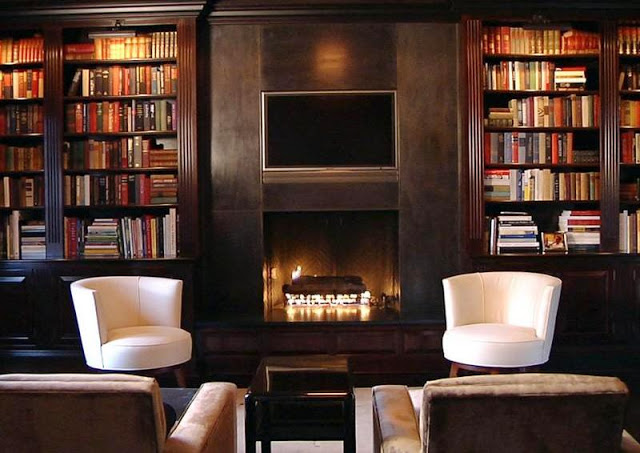dark wood paneled library in a mansion with a fireplace, four armchairs, two white, two brown, built in book shelves full of books and a wall mounted tv