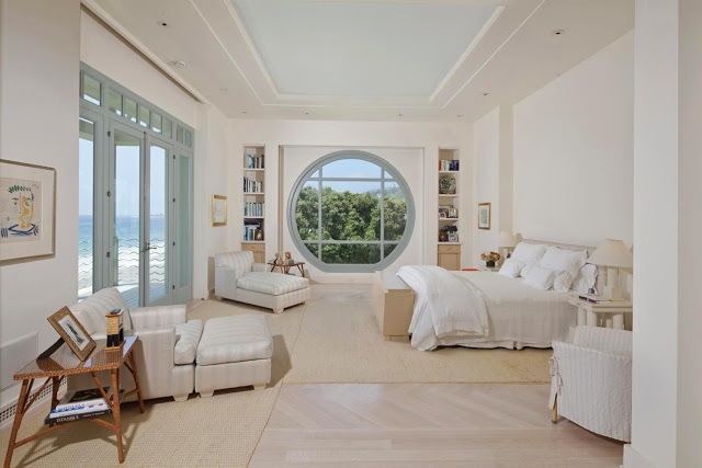 master bedroom with ocean view, blue accented circle shaped window and matching glass door, two ottomans in the corner and wicker chairs