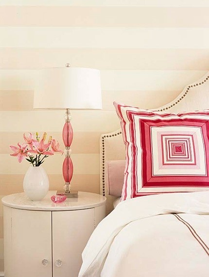 Bedroom with white upholstered headboard, a round night stand, pink and white lamp with a glass base, a graphic pillow and striped wall