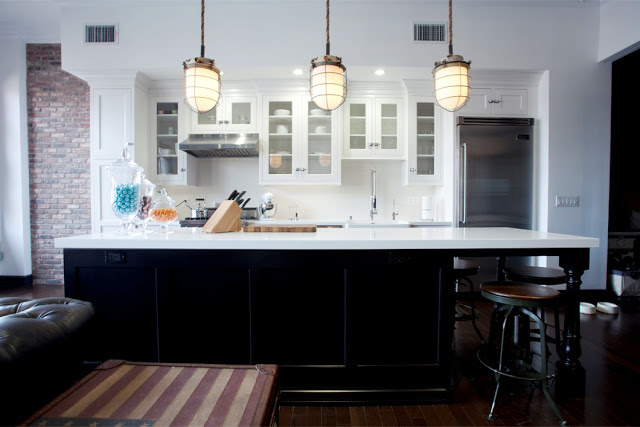 Kitchen in Daniel Lowe's Hollywood loft with three large pendant lights, dark black island, quartz counter tops, white kitchen cabinets, bar stools, wood florring, and a brick wall