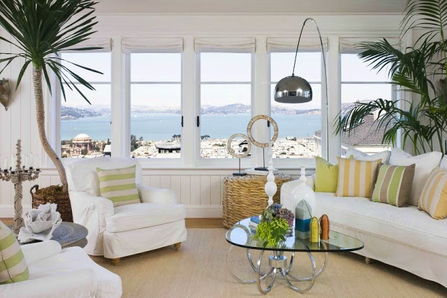 Family room in a san francisco mansion with white paneled walls, white sofa, two white armchairs with striped green and grey accent pillows, a glass coffee table, two palm trees, a modern metal reading lamp, and five windows with white rollup shades and an ocean view