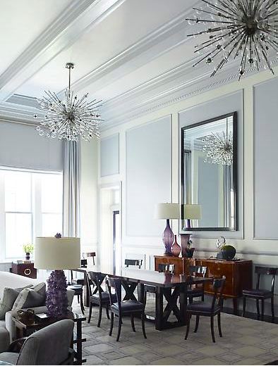 Dining room by Steven Gambrel with high coffered ceilings and decorative molding, two modern chandelier, grey-lavender painted walls to created a panel effect, floor length curtains, a greyish lavender area rug covered in squares.