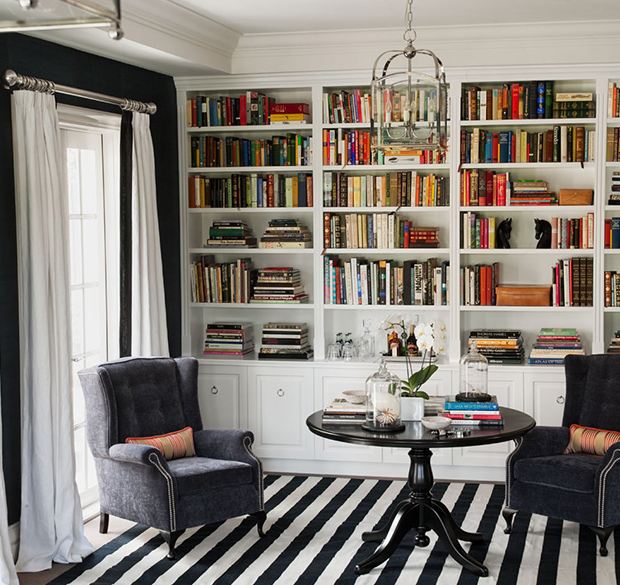 library with white built in bookshelves, black and white striped rug, velvet wing back armchairs, round wooden table, black wall with white floor length curtains