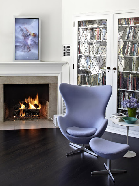 Library with dark wood floor, fireplace with white moulded mantel, a  lavender Arne Jacobsen's 1958 Egg Chair with matching foot rest, a white round side table and bookshelves with glass doors