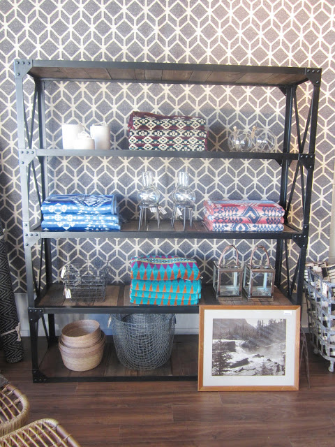 A metal shelf holding small objects propped in front of a graphic patterned flat weave dhurrie wool rugs are displayed hanging on the store's walls like wallpaper