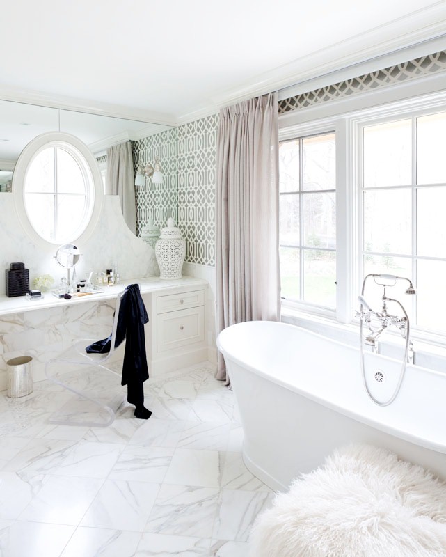 Girly bathroom with white marble floor, stand alone bathtub, Trellis wallpaper, vanity with a lucite chair under an oval window