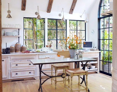 Kitchen with black encasemenet windows, exposed beams, silver pendant lights, cream drawers with black drawer pulls, a table with iron legs and two stools
