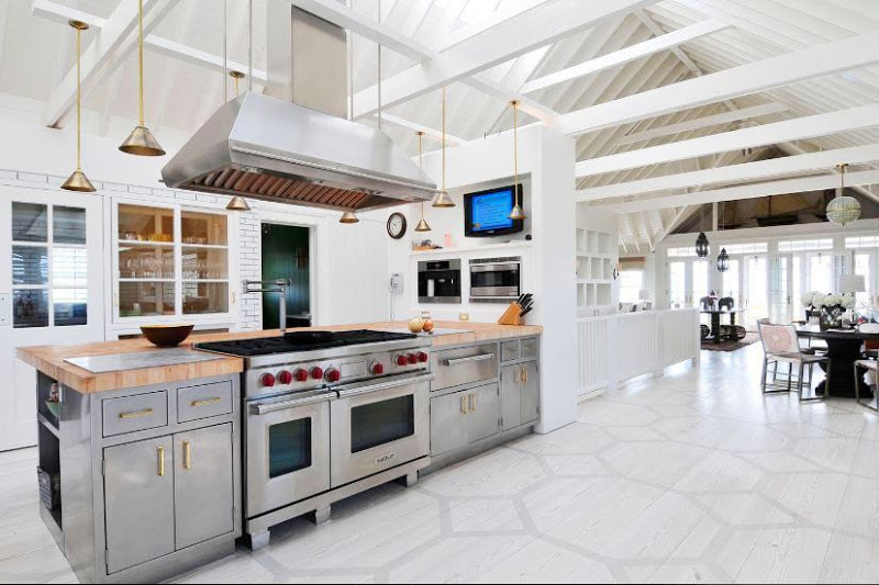 Huge kitchen in Sagaponack with high ceiling and exposed beams, state of the art appliances, stainless steel lower cabinets, brass pulls, brass pendant lights and a graphic painted floor