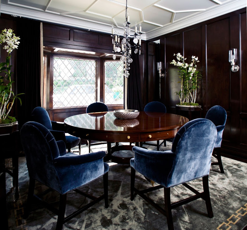 Dining room with white coffered ceiling, dark paneled walls, blue velvet armchairs around a wood table, a grey velvet rug and a crystal chandelier 