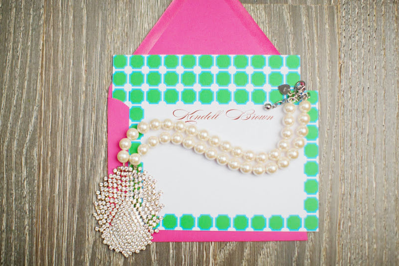White card with green and turquoise design, a hot pink envelopes and a pearl necklace 