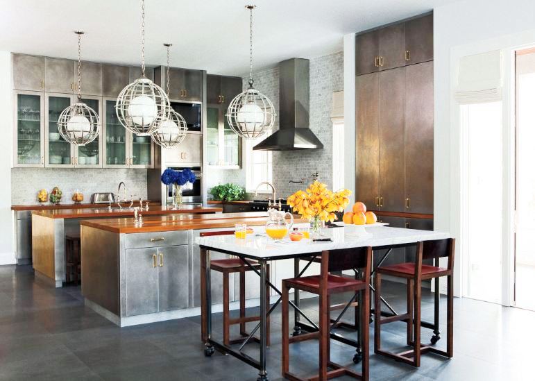 Kitchen with huge charcoal grey field tiles, stainless cabinets with brass pulls, teak countertops, four round pendant light caged, a wheeled table with white countertop surrounded by four bar chairs