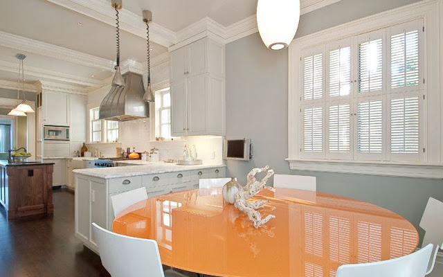 Breakfast nook with round orange table surrounded by white chairs, dark wood floor, grey walls, pendant lights, white countertops with matching cabinets and a wood island