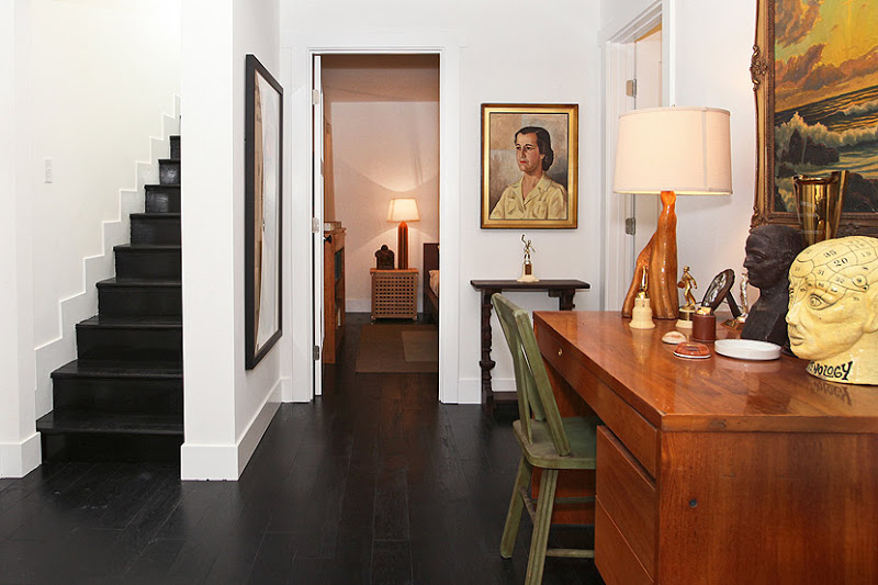 Foyer with stained wood floor, black stairs, a wood desk with busts on it and a natural wood lamp and at the desk is a green wood chair
