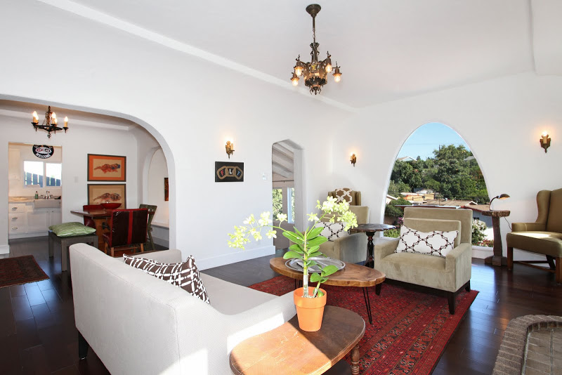 Open living plan in a Spanish Style home with an arched picture window, club chairs and a grey sofa with cococozy pillows and throws, dark wood floors, a red Moroccan rug, a brick fireplace and a chandelier