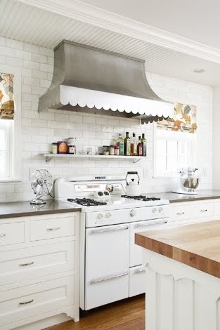 close up of rehkamp larson architects' vintage kitchen with a roper stove, a scalloped metal hood with subway tiles from counter to ceiling in the backsplash, the countertops are black and the cabinets under them are white.