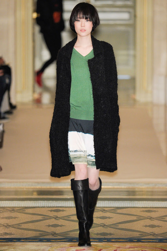 model from Agnes B.'s Fall 2011 Ready to Wear show. She's wearing knee high black boots, a slightly oversized green sweater, a white skirt with green stripes at the bottom and a long black jacket