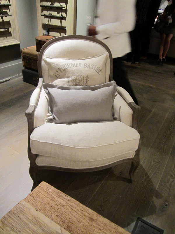 Taupe upholstered armchair with a large accent pillow that say "Ets Fournier Bastide" and a small grey rectangular pillow on a painted wood floor
