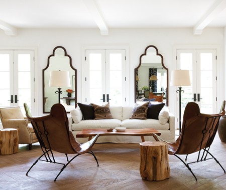 Living room with French doors, visible beams, Moorish floor mirrors, two leather butterfly chairs, reclaimed wood stools, light colored herringbone wood floors and a white upholstered sofa