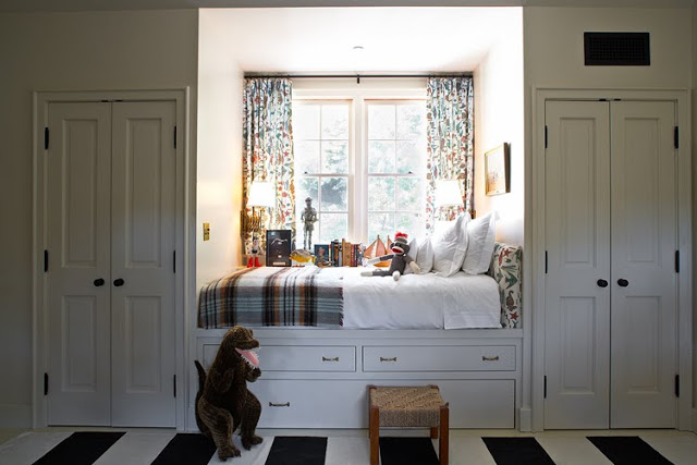 Built in window bed with white under bed drawers doubles as a window seat and reading area in a white bedroom with a black and white stripped rug, 