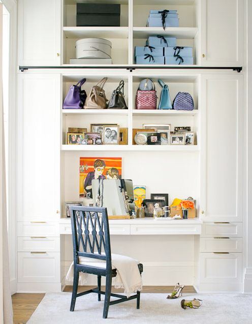 white built in desk/vanity in a walk in closet with a blue wooden chair. The shelves above the desk hold pictures, handbags and decorative boxes