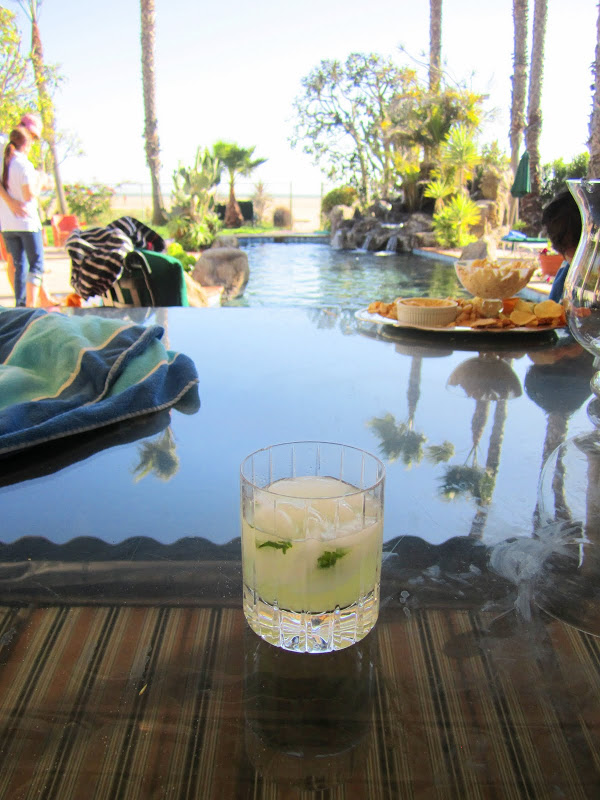 Mojito on a polished table reflecting palm trees and a striped canopy, in the distance there is a gorgeous pool surrounded by plants. Just behind them is a beach and an ocean view