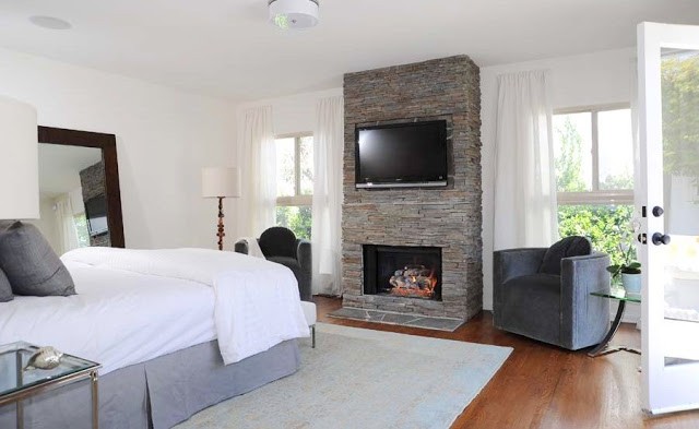 Master bedroom with a stone fireplace with a wall mounted flat screen TV, on each side of the fireplace is a grey armchair in front of a window with floor length white curtains, the room has wood floors, a grey area rug and a large mirror leans up against the wall. French doors lead outside. 
