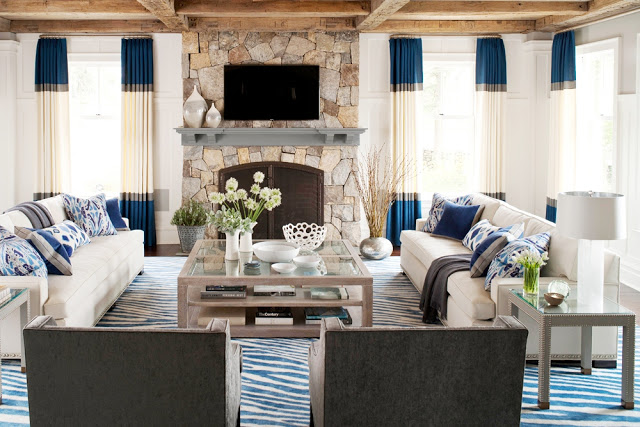 Living room with color block curtains, a stone fireplace, dueling facing sofas, exposed beam ceilings, and a large coffee table