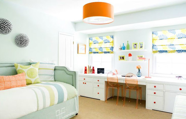 bedroom with an orange pendant light, mint green upholstered futon, wall length white desk with orange wire chairs with white cabinets with orange drawer pulls