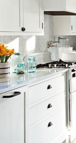 Close in a white kitchen with white drawers and cabinets, black drawer pulls, marble counter tops with blue glass bottles and a vase holding daffodils