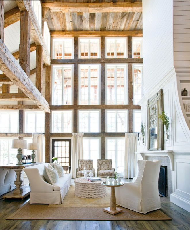 Living room with reclaimed wood beams, a super tall white wood paneled and plank covered fireplace, 2.5 story windows, a wood floor with a sea grass rug, a white sofa with matching armchair, and a round ottoman