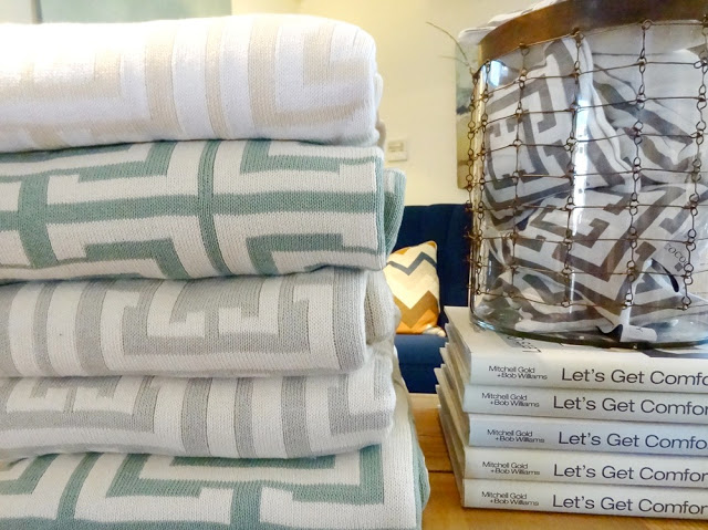 Five COCOCOZY throws are stacked on top of each other, to their right is a stack of books on top of which is a container holding COCOCOZY Lavender Sachets