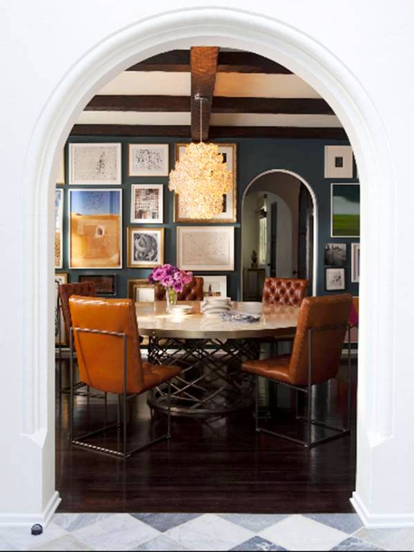 Arched entry way into the dining room. The dining room has a wood floor, round table on a metal base surrounded by tufted leather chairs, the walls are blue and covered from top to bottom in framed art, on the ceiling there is a modern chandelier
