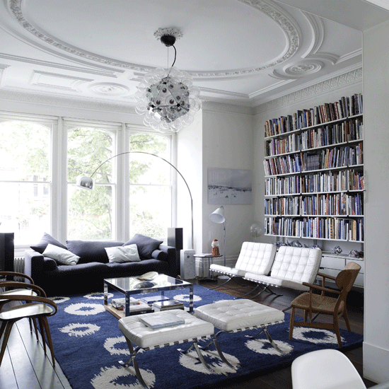 Living room with built in book shelves, traditional decorative ceiling molding, blue and white ikat rug, white barcelona chairs with matching ottomans, bentwood chairs, a modern light blub chandelier and a blue sofa