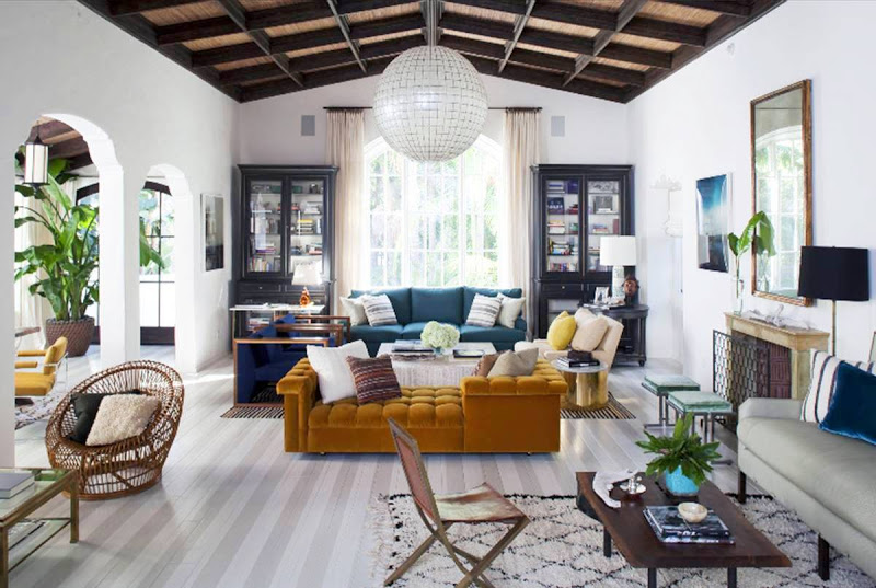 Living room with high ceiling, exposed beams, a round pendant light, striped floor, a blue sofa, black bookshelves on each side of an arched window, a tan chaise lounge and a fireplace