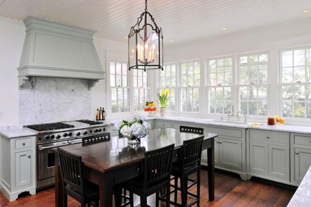 Alternative view of the kitchen's grey cabinets, marble counter tops, casement windows, stained oak table dining room table surrounded by matching chairs and a chandelier