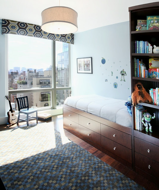 twin bed with drawers and a bookshelf attached, wood floor with rug covered in multicolored squares and a large window with aview of a city