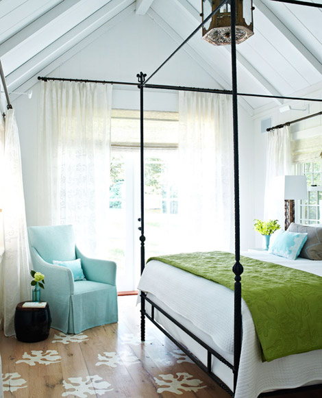 Bright bedroom with an iron canopy bed with white bedding and a bright green blanket draped across the foot of the bed, blue Holly Hunt armchair with a Leontine Linens boudoir pillow, painted reclaimed wood floors and breezy white floor length curtains