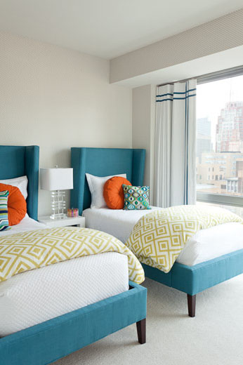 Clean and modern twin bedroom with white walls and floor, the turquoise upholstered beds have a wing back headboard, the orange accent pillows and a green and white down comforter folded across the bottom of the beds add a pop of color.