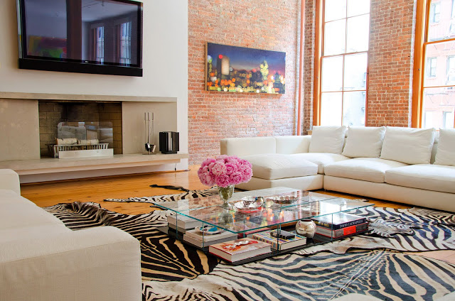 Alternative view of living room with the Cassina coffee table above. Here you can see the white sectional sofa, zebra print rugs, brick walls, fireplace, wall mounted TV and large windows.