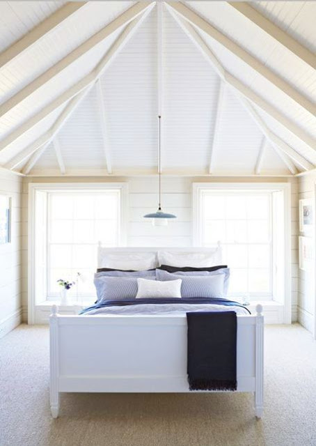 Simple, elegant bedroom with a bed with a white wooden frame and headboard, exposed beams, two windows and a single pendant light