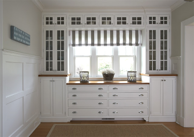 Butlers pantry with white cabinets with glass fronts, a striped roll up shade, wood floor and wall panel molding