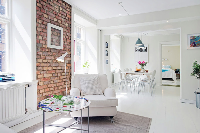 Alternative view of the living room's where you can see the painted, round coffee table, a white armchair, an exposed brick wall with an empty picture frame and the dining room 