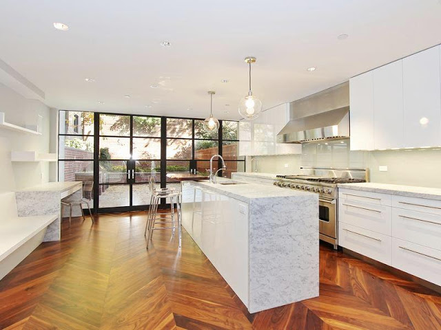 Sleek kitchen with herringbone wood floors, white drawers with long drawer pulls and cabinets, stainless appliances, pendant lights, an islance with marble countertop, and black encasement windows and door exiting to a patio