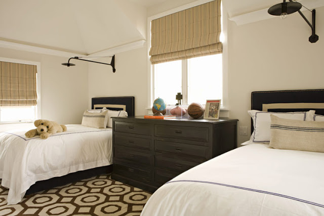 Twin bedroom with simple brown headboards with off white time, a brown and white graphic print rug, a dark wood chest of drawers, black wall mounted lights and roman shades on the window