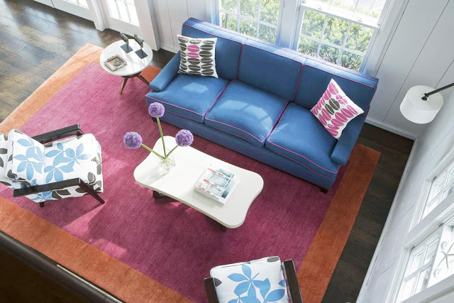 living room with stained wood floor, orange and purple area rug, blue sofa, white coffee table and accent table and two armchairs with white cushions with large blue and brown leaves printed on them