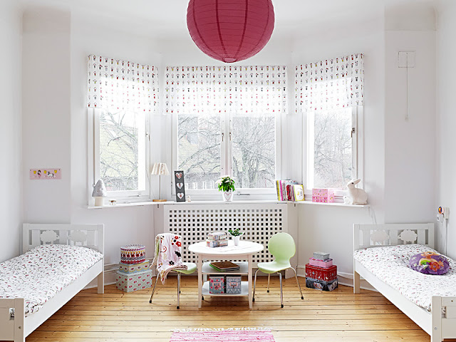 White children's bedroom with two beds with patterned bedding, pink spotted accents on the curtains, a small table with two green chairs and a large round pink lantern on the ceiling